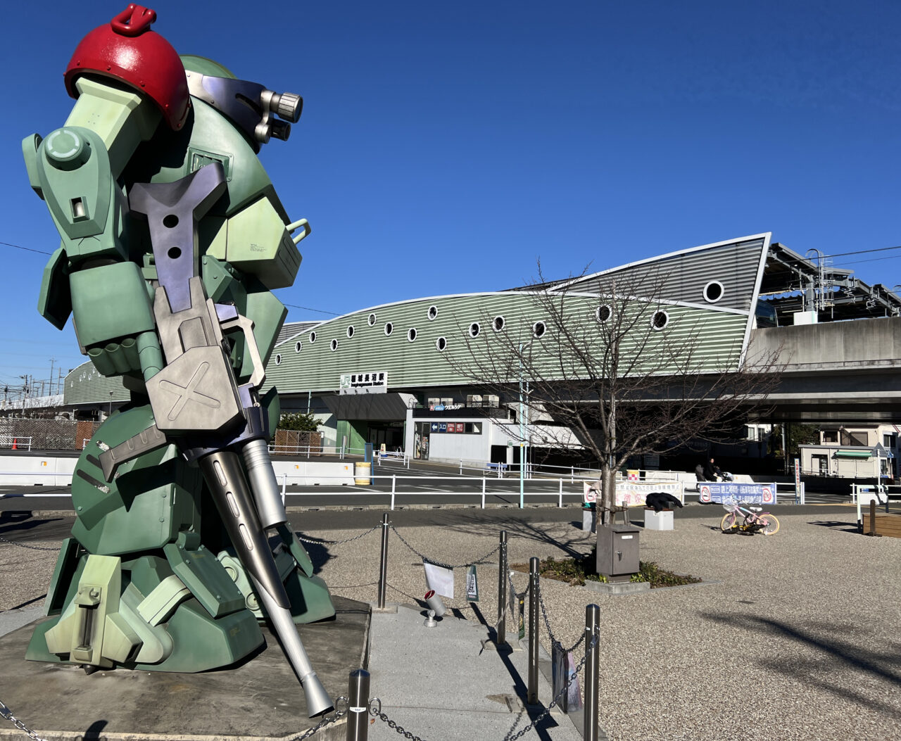 昼間のスコープドッグモニュメントと稲城長沼駅